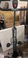 Front view of lightweight foldable Booty Gainer workout machine showing dual foot pedals, padded handlebars, and digital display on metal frame.