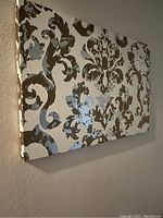 Side angle view of one canvas panel showing damask pattern in brown and gray on a white background, hung on beige wall.