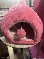 Front view of the pink plush cat house with an arched opening and hanging pom-pom toy inside, sitting on a small round table.