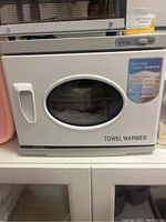 Front view of the white electric towel warmer with a small oval transparent window and control strip on top, door closed.