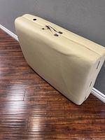 Closed massage table standing vertically showing beige exterior and carry handle.