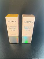 Photo showing two boxed Moira Glow Getter Hydrating Lip Oil products side by side, one in yellow-topped packaging and the other in white-topped packaging.