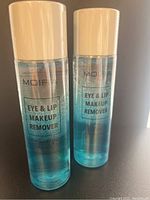 Two clear plastic bottles with off-white caps labeled Moira Eye & Lip Makeup Remover containing blue liquid, photographed front on with a dark background.