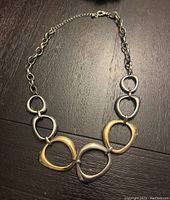 Silver necklace laid flat on dark surface, showing five large interlocking abstract links connected to smaller chain links and lobster clasp.