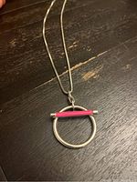 Photo of silver chain necklace with circular pendant and horizontal pink accent bar on a dark wooden surface.