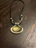 Photo shows silver necklace with trapezoidal pendant containing a multicolored stone, decorative silver beads on black cord, displayed on wooden surface.