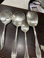 Photo showing four silver serving spoons of different sizes and styles arranged on a dark surface. One spoon has a slotted bowl, and three have solid bowls. Handles show varied designs and patterns.