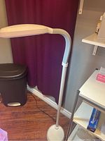 White adjustable arm standing lamp powered off, shown next to trash bin and shelving unit with purple curtain background