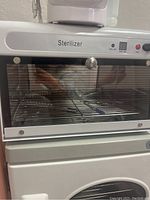 Front view of the sterilizer oven showing the control panel with digital timer and buttons, glass door, and interior wire racks.