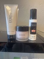 Three Sydoni skin care products, Vitamin C Cleanser, Rose Face Balm, and Rose Gelee Moisturizer on a black surface.