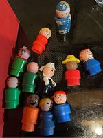 12 vintage Fisher Price plastic peg people figurines of various colors with some paint wear on a clear surface with black background.