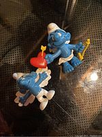 Photo showing two vintage Angel Smurf figures on reflective surface, both with white wings and white garments, one holding heart, one holding staff.