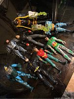 Seven Batman-related action figures laid out on dark surface, showing full bodies and poses, varying colors and designs representative of Batman and associated characters.