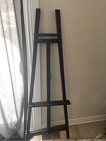 Photo showing a large black wooden easel folded and leaning against a wall near a window with curtains.