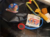 Photo of vintage Fisher Price medical kit items including black carrying case, toy stethoscope, bandage box, plastic syringe, and toy cup on dark surface.