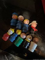Nine vintage Fisher-Price Little People plastic figurines laid on a dark surface, showing various characters with different colored outfits and hats.
