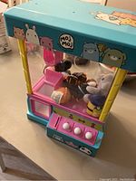 Mini claw machine showing blue body with cartoon characters, transparent inside with small plush toys visible, three control buttons on front.