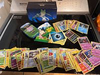 Photo showing a stack of about 90 Pokémon cards spread out, including energy cards and character cards, with two Pokémon-themed storage tins on a stove.