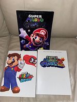 Three Super Mario video game guidebooks placed on a couch: Super Mario Galaxy Premium Edition, Super Mario Odyssey, and Super Mario Galaxy 2 Collector's Edition.