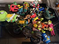 Various Teenage Mutant Ninja Turtles and related action figures scattered on black surface, showing multiple characters and a toy motorcycle.
