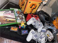 Photo shows various video game controllers, game cases, game cartridges, and a collectible figure arranged on a black surface.
