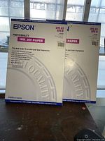 Two unopened Epson photo quality ink jet paper boxes with visible text and labels.