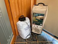 Photo showing two air beds with storage bags and a black electric pump on the floor next to a wooden wall and a door.