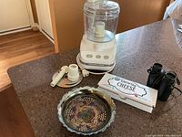 Overall view of KitchenAid food processor alongside carnival glass candy dish, mini binoculars, and cheese marker set on kitchen counter.