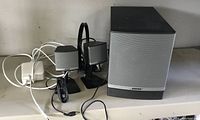 Photo of two small Bose satellite speakers on a stand and one large Bose subwoofer on a table, showing wired connections