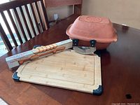 Photo showing terracotta Claybaker on table with lid, bamboo bread cutting board with black corner protectors, fiddle bow bread knife in original box, and Ulu chopping board knife.