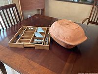 Photo showing terracotta Schlemmertopf claybaker next to bamboo drawer organizer containing several kitchen utensils on a wooden dining table.