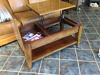 Maple coffee table with lift-top partially raised revealing storage compartment inside. Table legs and shelf visible. Shows wood grain and condition with chip.