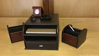 Desk accessories displayed on wooden surface including folding alarm clock in wood case, black stapler, magnetic paper clip holder, two rotating pen holders, and paper filing organizer with slots and drawer.