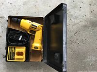 Yellow DeWalt corded electric drill and charger inside an open black metal storage case on concrete floor.