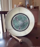 Front view of a round bodhran drum with white hide and colorful Celtic knots painted in green and black at the center, wooden drum stick lying in front.