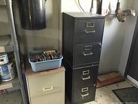 Photo showing three filing cabinets: one tall black 4-drawer metal cabinet, one beige 2-drawer cabinet, one cream 2-drawer cabinet. Small plastic tub with mixed CDs sits on top of beige cabinet.