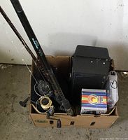 Top-down view of three fishing rods with attached reels packed in a cardboard box beside a black tackle box containing various fishing accessories.