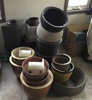 Large assortment of plant pots stacked and nested. Includes terracotta ceramic pots with drainage holes, black and beige plastic pots with textured patterns, square plastic pots, and glazed ceramic pots in yellow and brown tones. The pots vary in size from small to large and are used with some signs of wear.