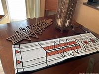 All items displayed on a wooden table: a geometric wooden wall hanger, a textile runner featuring geometric and wheat motifs, and a standing ambient side table light with glass panels and metal or wood framing.