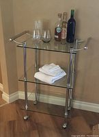 Glass serving cart with three glass shelves and chrome frame, shown with glassware, bottles, and towels placed on it.