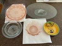 Four items on a counter: round pink Depression glass tray, clear pressed glass bowl, pink raised oval cookie tray, yellow ceramic olive serving dish with small jars