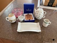 Photo of the full collection of glassware and barware items on a countertop including creamer, teapot, cups, tray, pink glass bowl, and Haig Club flask in original box.