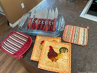 Photo showing 3 stacked Pyrex glass pans, 8 Paderno steak knives with wooden handles placed inside top pan, and 5 assorted hot pads beside them on counter