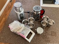 Image showing Delphino electric coffee grinder, mandolin slicer with extra blades in plastic bag, stainless steel and red Nespresso milk frother, six aluminium Nespresso cups and one white porcelain espresso cup on brown countertop.