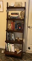 Full view of the dark wood 5 shelf ladder bookcase in a room, with books and decorative items on each shelf for scale, items are not included in sale.