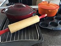 Photo shows orange Lagostina cast iron Dutch oven with lid, red KitchenAid cast iron lidded pot, black Wilton cookie pan, metal cooling rack, wooden rolling pin with red handles, and Paderno muffin tin.