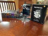 Black Nespresso single-serve coffee maker, boxed Aeroccino4 frother and multiple tubes of coffee pods displayed on wooden table.
