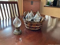 Five clear teardrop-shaped glass oil burners with metal tops and detailed metal bases placed on a wooden table. Four oil burners arranged in a woven basket and one standing outside the basket.