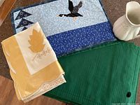 Photo showing yellow and white embroidered tablecloth, blue bird appliqué quilted tablecloth, six forest green pleated placemats partly visible beneath, and white ceramic pitcher.