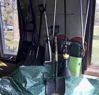 Photo showing assorted garden and outdoor tools including spades, rakes, snow scoops, and weed control sprayer against a window background with natural light.